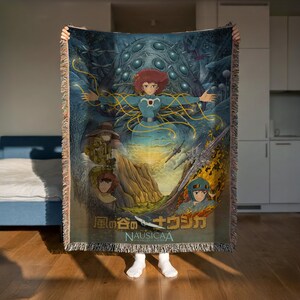 A person is holding up a colorful tapestry or blanket featuring an illustration of characters from the animated film "Nausicaa".