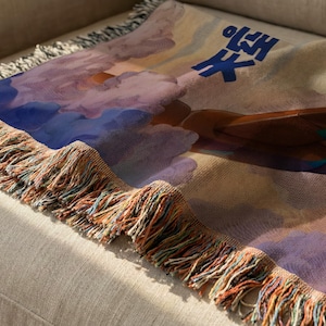 a colorful, abstract design with a mix of blue, purple, and orange colors, along with a cloud-like shape. The design appears to be a textile or fabric item, possibly a blanket or throw, with a fringed edge.