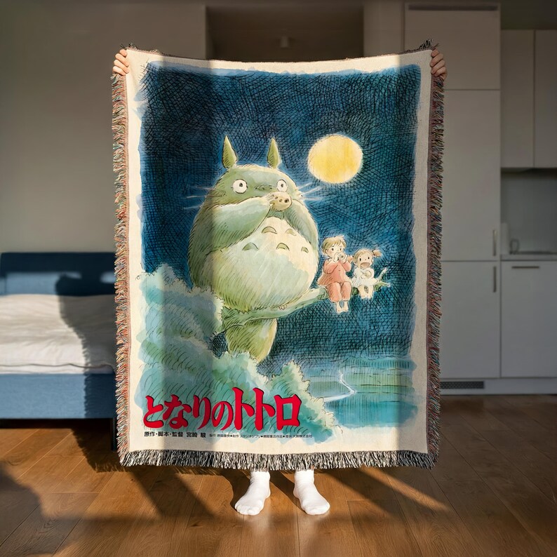 A person is holding up a large blanket featuring an illustration of a cat-like creature with a yellow moon in the background.