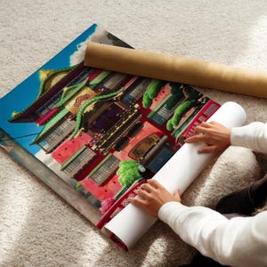 A person is sitting on the floor and rolling up a large poster of a colorful Asian building.