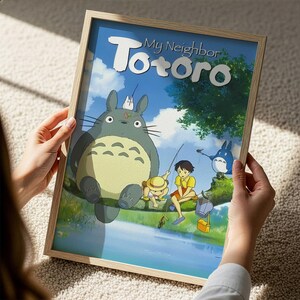 A person is holding a framed poster of the animated film "My Neighbor Totoro". The poster features the characters Totoro, a cat-like creature, and his friends in a forest setting.