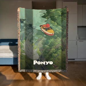 A person is holding up a large, colorful blanket with an illustration of a character riding a boat in a green underwater scene.