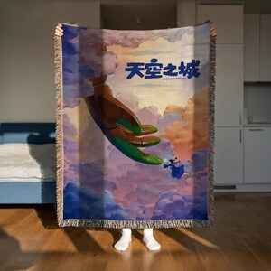 A person is holding up a colorful tapestry or blanket with a painting of a hand reaching out towards a person in the sky.