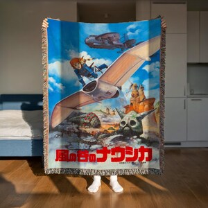 A person is holding up a colorful woven blanket featuring an illustration of a character and various fantastical elements, including a flying creature and a skull-like structure.
