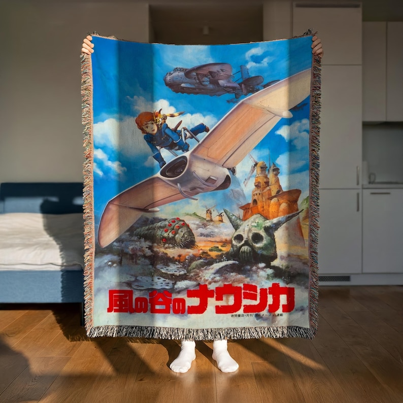 A person is holding up a colorful woven blanket featuring an illustration of a character and various fantastical elements, including a flying creature and a skull-like structure.