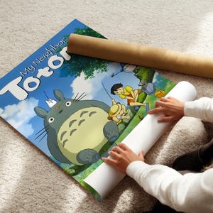 A person is sitting on the floor and placing a roll of paper under a poster featuring characters from the animated film "My Neighbor Totoro".