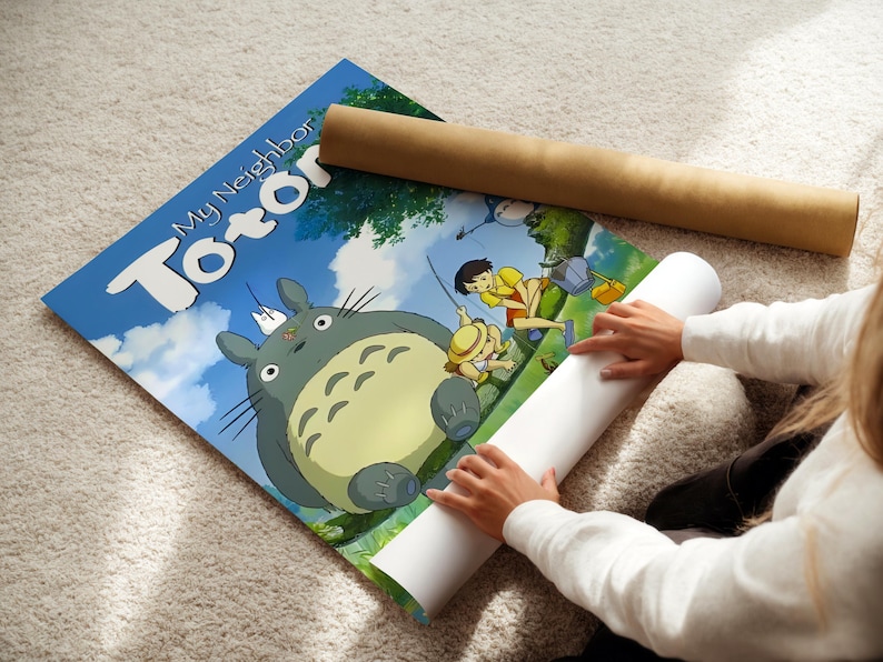 A person is sitting on the floor and placing a roll of paper under a poster featuring characters from the animated film "My Neighbor Totoro".