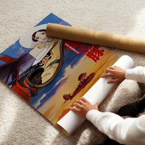 A person is sitting on the floor and placing a roll of paper under a poster featuring an illustration of a character and a scene with a red airplane.