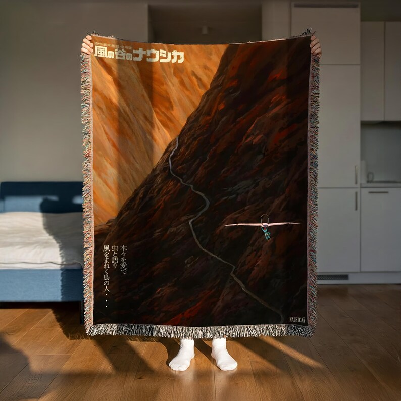A person is holding up a large tapestry or blanket with a mountain landscape design on it, standing in a room with a blue bed and white cabinets in the background.