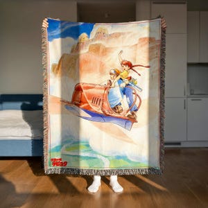 A person is holding up a colorful woven blanket with an illustration of two people riding a boat on a desert landscape.
