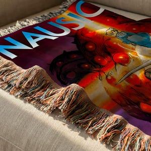 a colorful, abstract design with a mix of red, orange, and purple colors, along with various shapes and patterns. The design appears to be a textile or fabric item, possibly a blanket or throw, with a fringe or fringe-like texture.