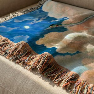 a close-up view of a colorful blanket with a painting of a blue sky and clouds on it, resting on a beige couch.