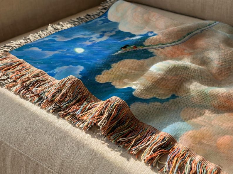 a close-up view of a colorful blanket with a painting of a blue sky and clouds on it, resting on a beige couch.