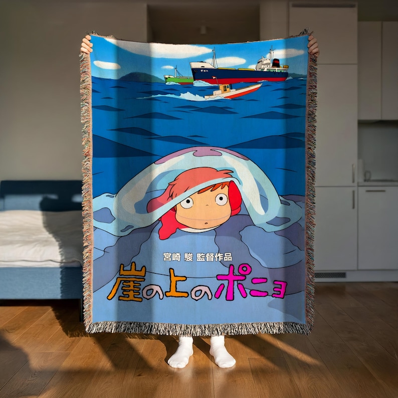 A person is holding up a colorful blanket featuring an illustration of Ponyo, a character from the animated film "Ponyo". The blanket has a large image of Ponyo peeking out from under a ship, and there is Japanese text on the blanket.