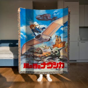 A person is holding up a colorful woven blanket featuring an illustration of a character and various fantastical elements, including a flying creature and a skull-like structure.
