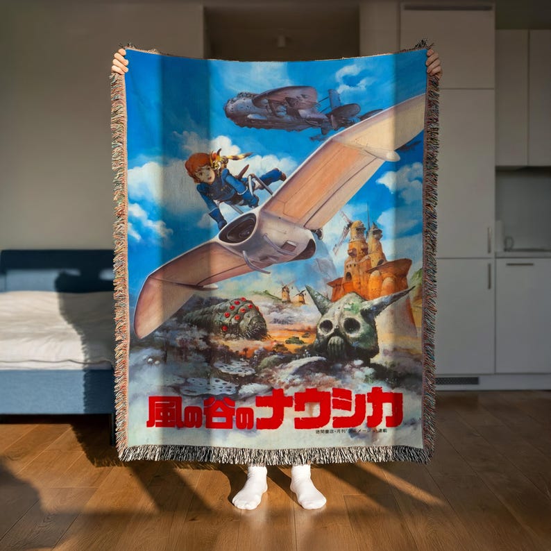 A person is holding up a colorful woven blanket featuring an illustration of a character and various fantastical elements, including a flying creature and a skull-like structure.