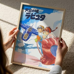 A person is holding a framed picture of an animated scene featuring a blue airplane and two characters, one of whom is wearing a helmet.