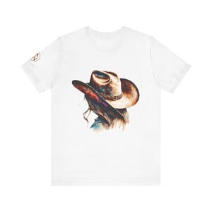May include: White t-shirt featuring a watercolor illustration of a person wearing a cowboy hat. The hat is brown with a decorative band. The artwork includes shades of brown, blue, and purple. The shirt has a crew neck and short sleeves.