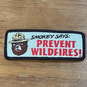 May include: Rectangular patch featuring Smokey Bear with the text "Smokey Says: Prevent Wildfires!" The patch has a black border, a light blue background, and the text and Smokey Bear are in red, brown, and white.