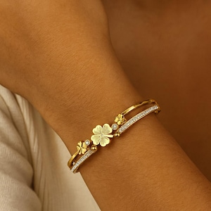 May include: Gold-toned bracelet featuring a four-leaf clover design with small, sparkling accents. The bracelet has a delicate, elegant appearance, with a double-band design. The clover is the focal point, with smaller floral details. The bracelet is worn on a wrist.