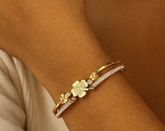 May include: Gold-toned bracelet featuring a four-leaf clover design with small, sparkling accents. The bracelet has a delicate, elegant appearance, with a double-band design. The clover is the focal point, with smaller floral details. The bracelet is worn on a wrist.