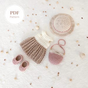 May include: A crochet doll outfit set in neutral tones, including a dress, hat, purse, and shoes. The dress is two-toned, with a cream-coloured bodice and a brown skirt. The hat and purse have pink accents. The set is displayed on a white surface.
