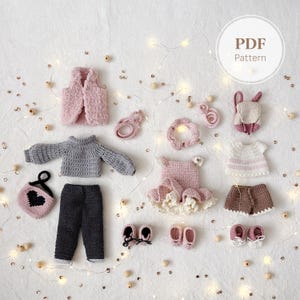 Könnte beinhalten: Eine Sammlung handgefertigter Häkelpuppenkleidung in verschiedenen Stilen und Farben. Das Set enthält eine rosa Weste, einen grauen Pullover, eine schwarze Hose, eine Handtasche, ein Kleid, Shorts, Schuhe und Accessoires. Die Artikel sind auf einer weißen Oberfläche mit dekorativen Lichtern angeordnet.