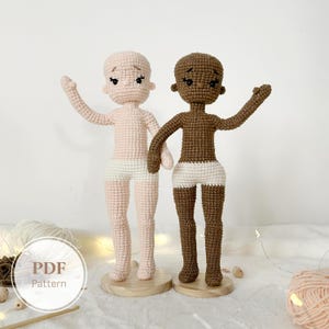 Puede incluir: Dos muñecas de ganchillo, una rosa claro y otra marrón, están una al lado de la otra. Cada muñeca tiene una expresión neutra, pantalones cortos blancos y los brazos levantados. Las muñecas están sobre bases de madera. La imagen incluye un gráfico circular con el texto "PDF Pattern".