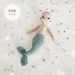 May include: A crocheted mermaid doll with a light pink torso and a blue-green tail. The doll has small black eyes and is lying on a white surface. The image includes the text "PDF Pattern" in a white circle.