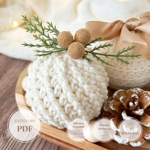 May include: A cream-colored crocheted sphere ornament topped with three gold textured berries and green pine sprigs. The ornament features a thick yarn texture and is accompanied by a pinecone and a beige ribbon bow.