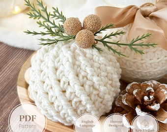 May include: A round, cream-colored crocheted ornament featuring a textured knit pattern. It is topped with three small, gold-colored textured spheres and green faux pine sprigs. A beige satin ribbon bow is attached to the side. Text reads PDF Pattern.