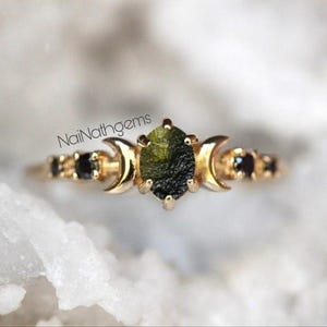 May include: A gold ring featuring a central oval-shaped green gemstone, flanked by crescent moon designs and black gemstones. The ring has a delicate band and is set against a soft, blurred background. The text "NaiNathgems" is visible on the image.