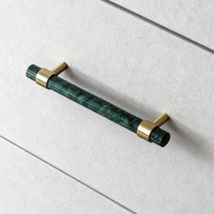 May include: Close-up of two cabinet drawer pulls. Each pull has a cylindrical, dark green marble-effect handle with gold-coloured metal brackets. The pulls are mounted on a light grey cabinet.