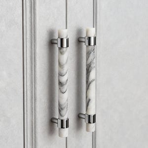 May include: Two cabinet pulls with a white and gray marble design. The pulls have a cylindrical shape with silver-colored metal accents. The cabinet doors are a light gray color.