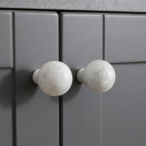 Könnte beinhalten: Nahaufnahme eines grauen Schranks mit zwei weißen Marmorknöpfen. Die Knöpfe sind rund und haben ein gesprenkeltes Muster. Die Schranktüren sind einfarbig grau.
