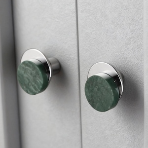 Op de afbeelding: Close-up van twee kastknoppen. Elke knop heeft een cirkelvormige, groene marmeren steen op een zilverkleurige basis. De knoppen zijn gemonteerd op een lichtgrijze kastdeur, wat een moderne esthetiek creëert.