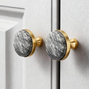 May include: Two cabinet knobs with a round, gray and white marbled design, accented by a gold-colored metal ring and base. The knobs are mounted on a white cabinet door, offering a modern aesthetic.
