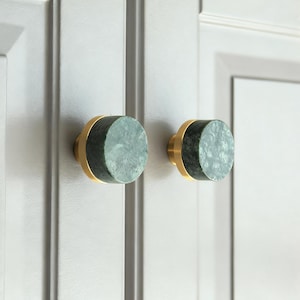 Può includere: Due pomelli per mobili con un centro circolare in pietra verde maculata e una base color oro. I pomelli sono montati su un'anta di armadio grigio chiaro. I pomelli sono un elemento decorativo per l'hardware.
