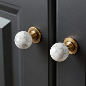 Op de afbeelding: Close-up van twee kastknoppen. Elke knop heeft een witte marmeren bol met grijze adering, geplaatst op een messingkleurige basis. De knoppen zijn gemonteerd op een donkergrijze kastdeur, wat een contrast in kleur en textuur creëert.