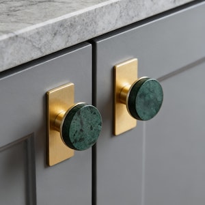 May include: Close-up of two cabinet doors with decorative hardware. Each door features a rectangular brass backplate with a round, green marble knob. The cabinet doors are painted a muted gray, and the countertop is a light gray marble.