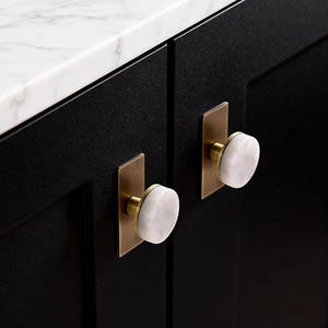 May include: Close-up of two cabinet doors with white marble knobs and brass rectangular backplates. The doors are black, and the countertop is white marble. The knobs have a rounded shape.