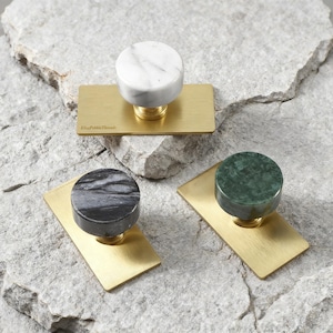 May include: Three circular cabinet knobs featuring polished stone tops in white, black, and green, each mounted on a rectangular brushed brass backplate. The white knob displays the text ElisePebbleThreads on its brass base.