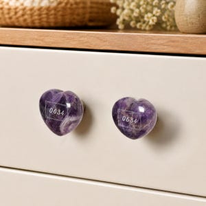 May include: Two heart-shaped cabinet knobs made of polished purple amethyst stone. Each knob features a rectangular white border containing the number 0634 printed in a clear, sans-serif font on the front surface of the stone.