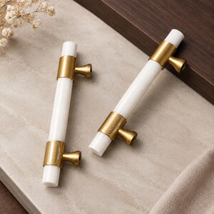 Peut inclure: Deux poignées de placard cylindriques composées d'un corps blanc lisse et de deux supports en métal doré brossé. Chaque support présente un bouton évasé et le mot VALENTIN gravé sur la surface. La quincaillerie a un design linéaire.