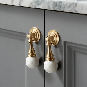 Könnte beinhalten: Zwei Schrankgriffe mit Messing-Finish und weißen Marmorkugeln. Die Griffe sind an einer grauen Schranktür montiert. Die Hardware hat ein klassisches, elegantes Design.