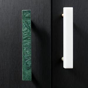 Puede incluir: Dos tiradores de armario, uno de mármol verde y otro blanco, sobre un fondo de madera oscura. El tirador verde tiene un patrón texturizado, mientras que el blanco es liso. Ambos son rectangulares y están montados con herrajes de latón.