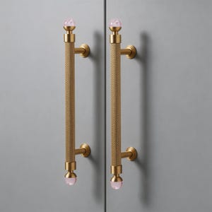 May include: Two gold-toned cabinet pulls with a textured, cylindrical design. Each pull features a pink, spherical accent at the top and bottom. The pulls are mounted on a light gray surface.