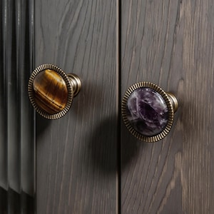 Puede incluir: Primer plano de dos tiradores de armario. Un tirador presenta una piedra de ojo de tigre con tonos marrones y dorados, y el otro tiene una piedra de amatista púrpura. Ambos tiradores tienen un borde y un vástago decorativos de metal color latón.