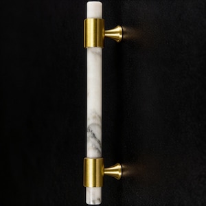 May include: A cabinet pull with a white marble centre and gold-toned metal accents. The vertical bar is approximately 15 cm long. The hardware is mounted on a black surface.