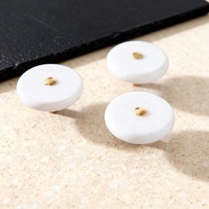 May include: Three white marble cabinet knobs with gold-colored accents. The knobs are round and have a polished finish. The hardware is designed for use on drawers or cabinets.